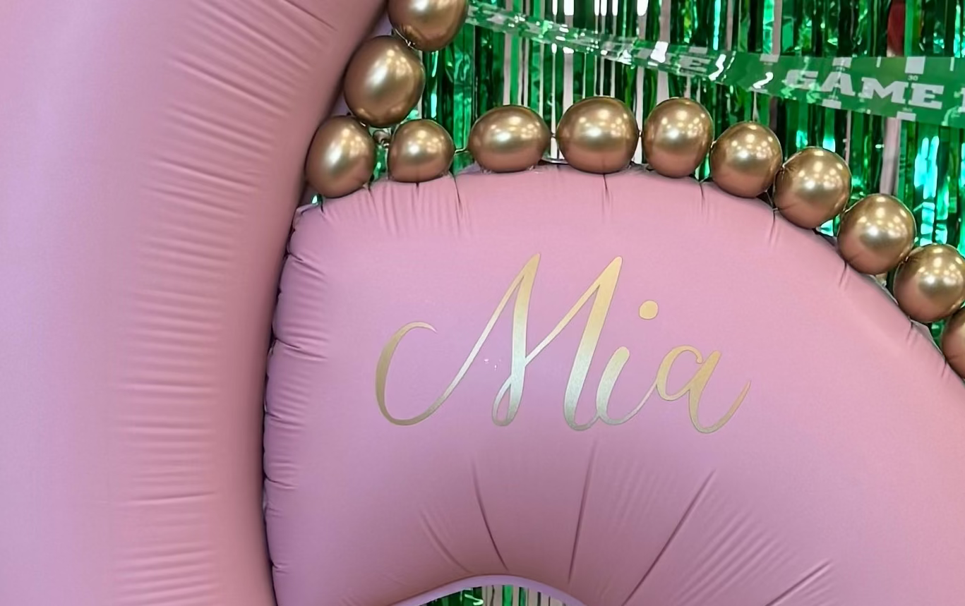 Pink balloon number '6' with gold beads and 'Mia' written on it, surrounded by colorful balloons and green tinsel.